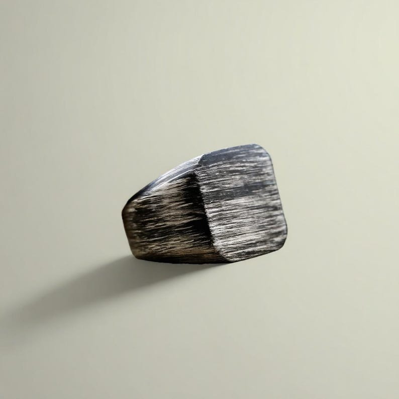 May include: A black and silver ring with a rectangular shape. The ring has a textured, brushed metal appearance. The ring is a statement piece, suitable for everyday wear.