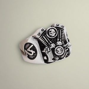 May include: A silver-toned ring featuring a detailed engine design with a skull and pistons. The ring has a textured finish and a side emblem with the letters "K" and "S" inside a circle. The ring is likely made of metal.