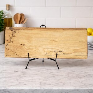 Peut inclure: Planche de service rectangulaire en bois avec un motif de grain naturel, présentée sur un support en métal noir. La planche est de couleur marron clair avec des marques sombres en forme d'éclair. Le support a une base à trois pieds.