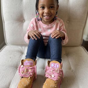 May include: Tan boots with pink accents and embellishments. The boots feature the word "EDITION" on one boot and "CEO" on the other. The boots have a studded design and pink laces.
