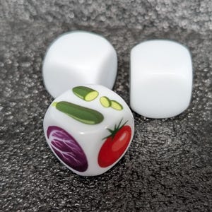 Pode incluir: Três dados brancos de seis lados. Um dado em primeiro plano apresenta ilustrações coloridas de um pepino verde, um tomate vermelho e um repolho roxo. Os outros dois dados são brancos lisos e posicionados atrás do dado ilustrado.