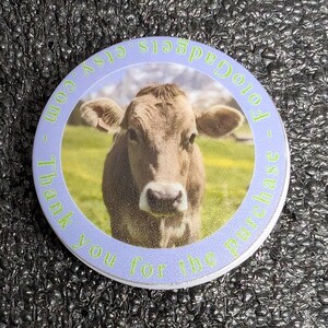 Op de afbeelding: Een ronde knoop met een lichtblauwe rand. Het toont een bruine koe in een groen veld. De tekst "FotoGadgets.etsy.com - Thank you for the purchase" is in het groen rond de rand gedrukt.