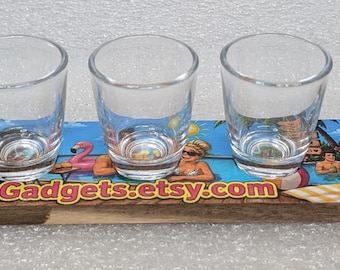 Op de afbeelding: Een houten serveerplank met een kleurrijke tropische strandscène en de tekst FotoGadgets.etsy.com, met zes heldere glazen shotglaasjes op een rij. De plank heeft een lang handvat met een gat aan het uiteinde om hem op te hangen.