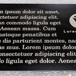 Op de afbeelding: Een zwarte rechthoekige kaart met witte tekst en een cirkelvormig logo. De tekst bevat de zin "Lorem ipsum dolor sit amet" en andere Latijnse tekst. Het logo toont twee gestileerde figuren. Het oppervlak van de kaart lijkt reflecterend.