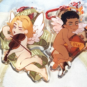 May include: Two clear acrylic charms depicting cherubic figures with wings. One plays a violin, the other holds a container with flames. Both have a banner with text. The charms have red and silver hardware.