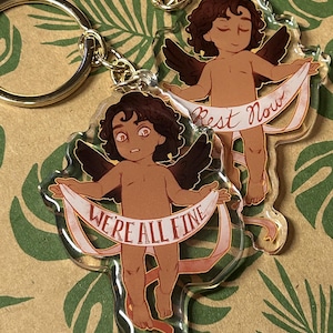 Może przedstawiać: Dwa przezroczyste akrylowe breloki w kształcie aniołów z brązowymi włosami i skrzydłami. Jeden anioł trzyma transparent z napisem "Rest Now", a drugi trzyma transparent z napisem "We're All Fine".