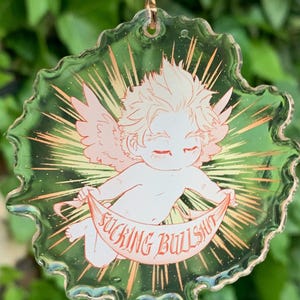 May include: A clear acrylic keychain featuring a cherubic figure with wings, holding a banner that reads "FUCKING BULLSHIT" in red text. The design has a green and gold background with a gold-colored chain.