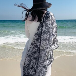 Peut inclure: Un chapeau de soleil noir avec un bord marron et une &eacute;charpe &agrave; motifs noir et blanc drap&eacute;e sur une robe blanche. La personne se tient sur une plage de sable, face &agrave; l'oc&eacute;an sous un ciel bleu clair.