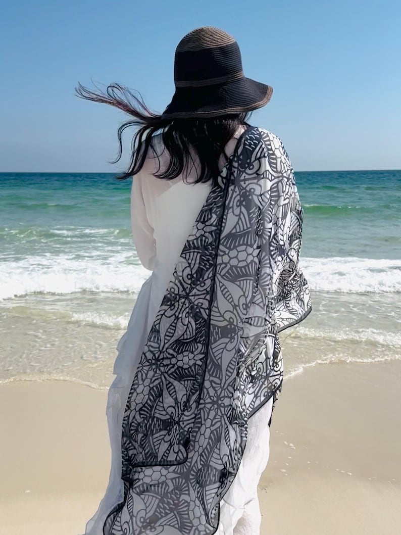 Peut inclure: Un chapeau de soleil noir avec un bord marron et une &eacute;charpe &agrave; motifs noir et blanc drap&eacute;e sur une robe blanche. La personne se tient sur une plage de sable, face &agrave; l'oc&eacute;an sous un ciel bleu clair.