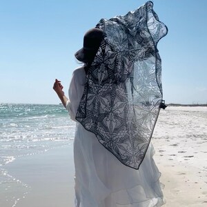 Peut inclure: Une personne portant un chapeau noir et une robe blanche se tient sur une plage, avec une &eacute;charpe &agrave; motifs noirs et blancs flottant dans le vent. L'oc&eacute;an et le ciel bleu sont visibles en arri&egrave;re-plan.