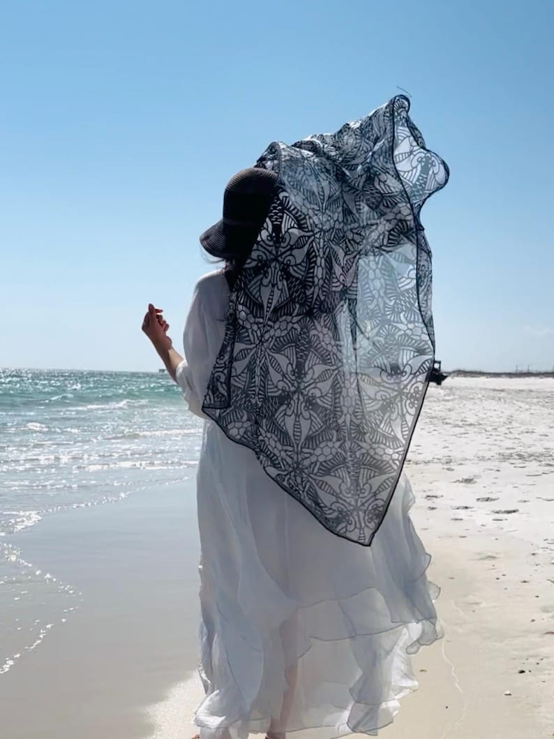 Peut inclure: Une personne portant un chapeau noir et une robe blanche se tient sur une plage, avec une &eacute;charpe &agrave; motifs noirs et blancs flottant dans le vent. L'oc&eacute;an et le ciel bleu sont visibles en arri&egrave;re-plan.