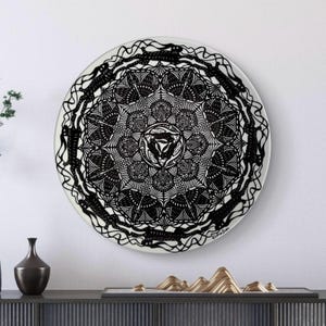 Könnte beinhalten: Eine runde, weiße dekorative Platte mit einem detaillierten schwarzen Mandala-Design. Das komplizierte Muster zeigt eine zentrale geometrische Form, umgeben von symmetrischen floralen und abstrakten Elementen. Die Platte ist an einer weißen Wand befestigt.
