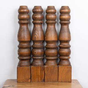 Könnte beinhalten: Vier Holzmöbelfüße mit warmbraunem Finish. Jeder Fuß hat eine quadratische Basis und ein gedrehtes, dekoratives Design mit abgerundeten Abschnitten. Die Holzmaserung ist sichtbar und verleiht Textur. Die Beine sind vertikal angeordnet.