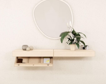 Puede incluir: Una estantería flotante blanca con un cajón, una planta en maceta y dos pequeños objetos decorativos. La estantería está montada en una pared blanca con un espejo blanco de forma orgánica encima.
