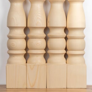 May include: Four unfinished wooden table legs. Each leg is light-colored, with a square base, a series of rounded and cylindrical sections, and a flared top. The legs are arranged in a row against a white background.