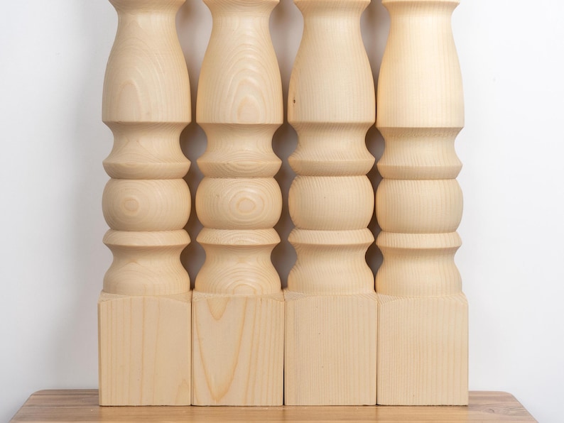May include: Four unfinished wooden table legs. Each leg is light-colored, with a square base, a series of rounded and cylindrical sections, and a flared top. The legs are arranged in a row against a white background.
