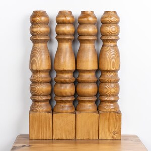 May include: Four wooden furniture legs with a light brown finish. Each leg has a square base and a turned, stacked design with rounded sections. The wood grain is visible, adding texture to the pieces.