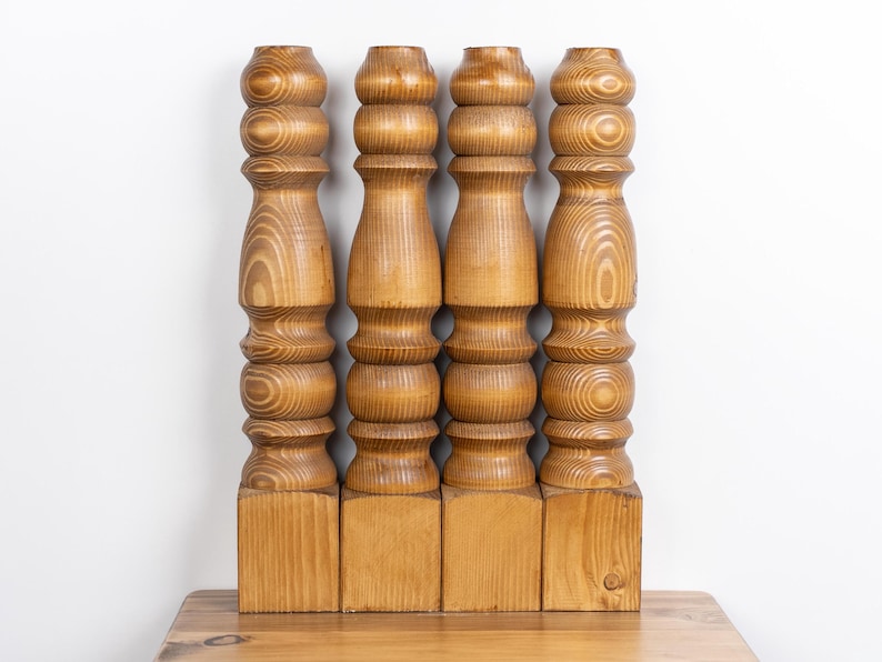 May include: Four wooden furniture legs with a light brown finish. Each leg has a square base and a turned, stacked design with rounded sections. The wood grain is visible, adding texture to the pieces.