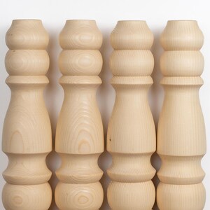 May include: Four unfinished wooden furniture legs. Each leg is light-colored, with a turned design featuring rounded sections and a wider base. The legs are arranged vertically against a white background, showcasing their shape and wood grain.