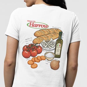 May include: White t-shirt featuring a food illustration with tomatoes, carrots, bread, olive oil, and an onion. The text "bisque à la Harrow" is above the illustration, with additional text labels.