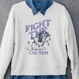 May include: White sweatshirt with the text "FIGHT THE CIS-TEM" in purple, featuring a graphic of a horse and rider. The sweatshirt is layered over a denim shirt. The design includes the year "1995".