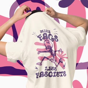 May include: Cream-colored t-shirt with a purple and pink graphic design. The design features a skeleton and the text "MORE FAGS LESS FASCISTS". The person is wearing blue jeans.
