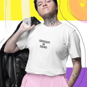 Può includere: Una maglietta bianca con la scritta "GENDER IS DEAD" è abbinata a pantaloni rosa e una giacca di pelle nera. Lo sfondo presenta un design astratto giallo, bianco, viola e nero con cerchi rosa e arancioni e pennellate blu.