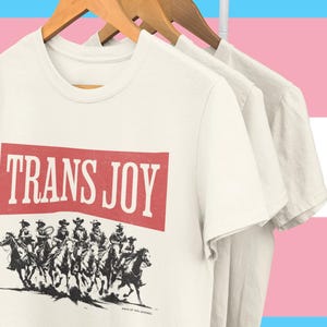 Op de afbeelding: Off-white T-shirts hangen aan houten kleerhangers. Het voorste shirt toont de tekst "TRANS JOY" in een rode rechthoek boven een zwart-wit illustratie van ruiters te paard. De achtergrond heeft de kleuren van de transgender pride vlag.