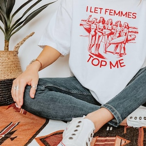 May include: White t-shirt with a red graphic of women surfing and the text "I Let Femmes Top Me".