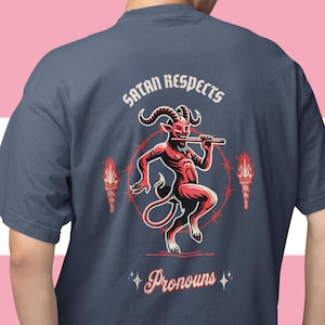 May include: A navy blue t-shirt featuring a red and black devil graphic with a flute, encircled by a design and torches. The text reads "Satan Respects Pronouns" above and below the image.