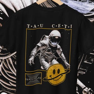 Puede incluir: Camiseta negra con un gráfico de un astronauta con un traje espacial cerca de un planeta amarillo con una cara sonriente. El texto "T.A.U. CETI" está impreso encima de la imagen del astronauta en un marco rectangular dorado.