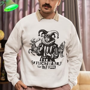 May include: White sweatshirt with a black and white graphic of a jester, deer, and wolf. The text "FITCHIVALRY and The Fool" is printed below the graphic. The person is wearing a gray cap and pink pants.