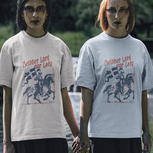 May include: Two people wearing light grey t-shirts with the text "Neither Lord nor Lady" and a graphic of a knight on horseback. They are holding hands, standing on grass near water.