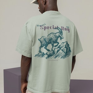 May include: A light green t-shirt with a graphic of a mountain goat on a rocky mountain. The text "Mommy's Special Boy" is above the goat in purple. The shirt is worn by a person wearing a beige baseball cap.