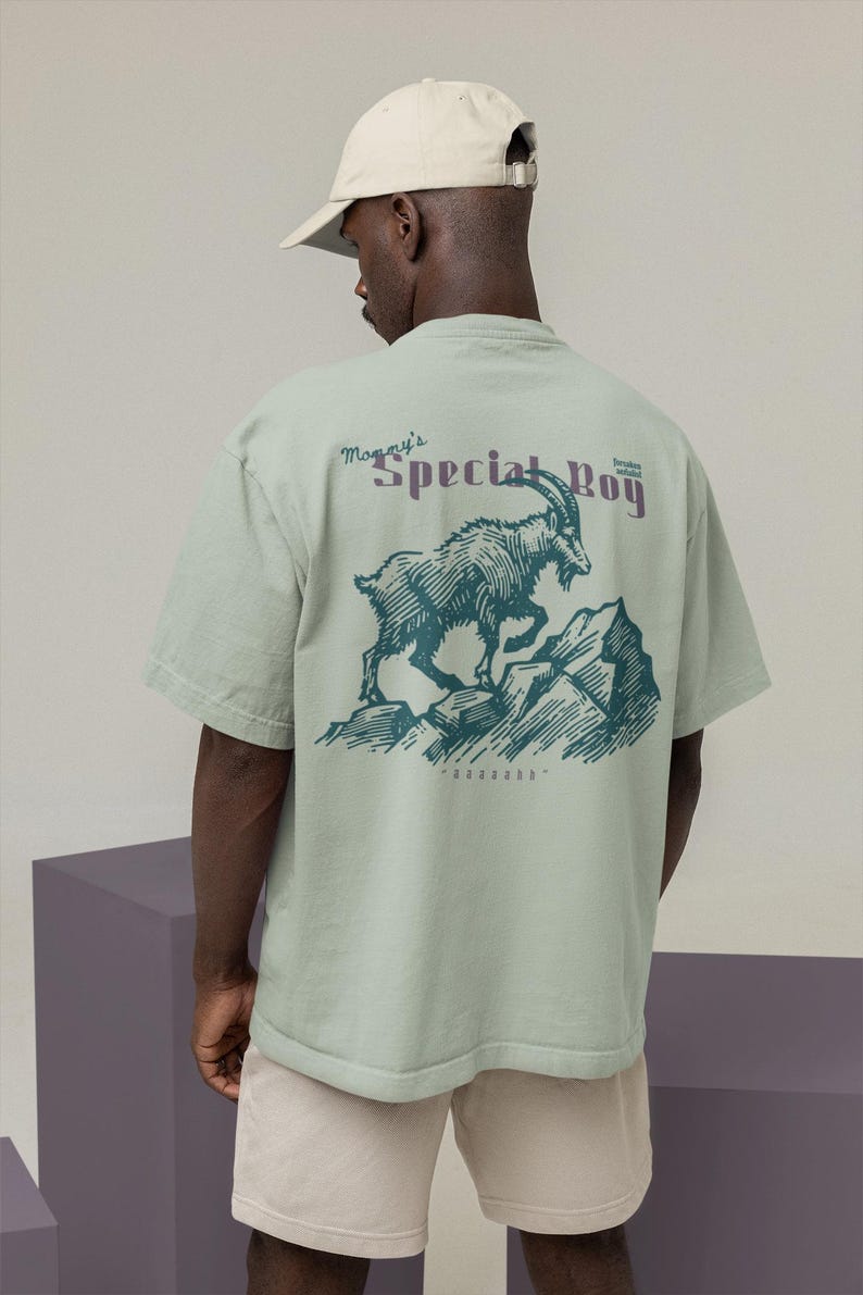 May include: A light green t-shirt with a graphic of a mountain goat on a rocky mountain. The text "Mommy's Special Boy" is above the goat in purple. The shirt is worn by a person wearing a beige baseball cap.