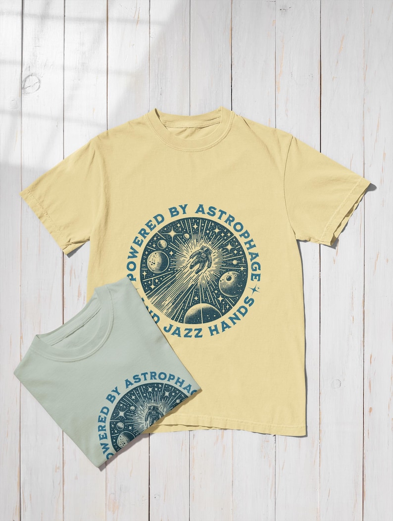 Może przedstawiać: Dwie koszulki z kr&oacute;tkim rękawem, jedna jasnoż&oacute;łta i jedna jasnoniebieska, z okrągłą grafiką. Grafika przedstawia astronautę, planety i tekst "POWERED BY ASTROPHAGE AND JAZZ HANDS" w ciemnozielonym kolorze.