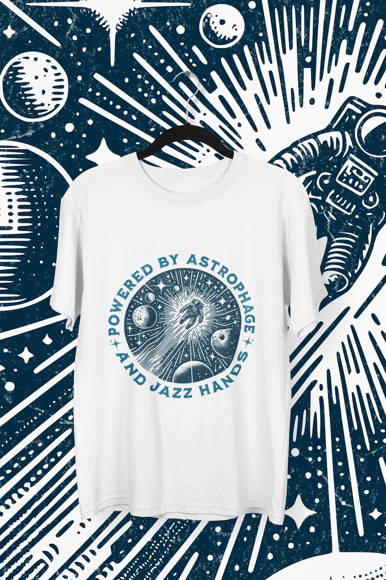 Może przedstawiać: Biała koszulka z okrągłą niebieską grafiką przedstawiającą astronautę, planety i napis "POWERED BY ASTROPHAGE AND JAZZ HANDS". Koszulka wisi na czarnym wieszaku na ciemnoniebieskim tle z niebiańskimi wzorami.