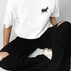 May include: White t-shirt with a black goat graphic and the words "prepotente pony" in red. The shirt is worn with black ripped jeans and white sneakers. The overall style is casual and modern.