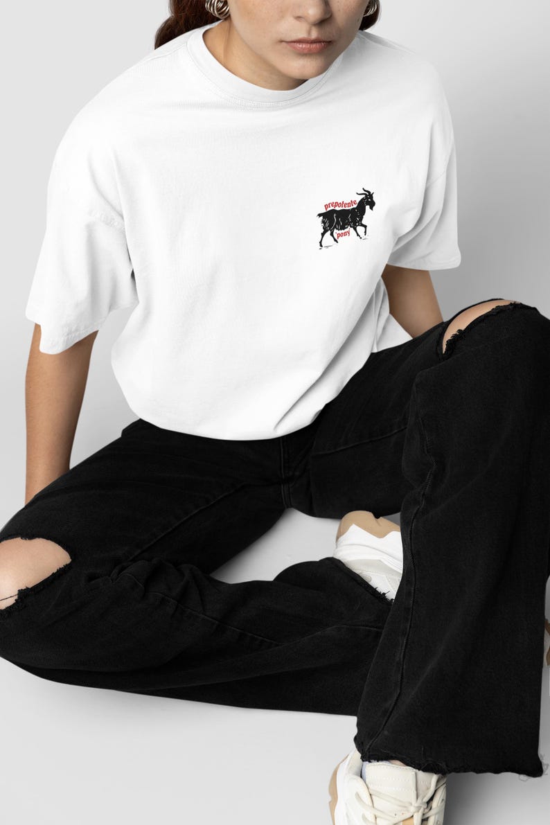 May include: White t-shirt with a black goat graphic and the words "prepotente pony" in red. The shirt is worn with black ripped jeans and white sneakers. The overall style is casual and modern.