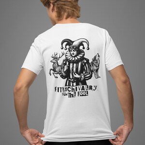 May include: White t-shirt with a black and white graphic of a jester figure, a deer, and a wolf. The text "FITUCHIVALRY and THE FOOL" is printed below the figures. The shirt is being worn by a person.