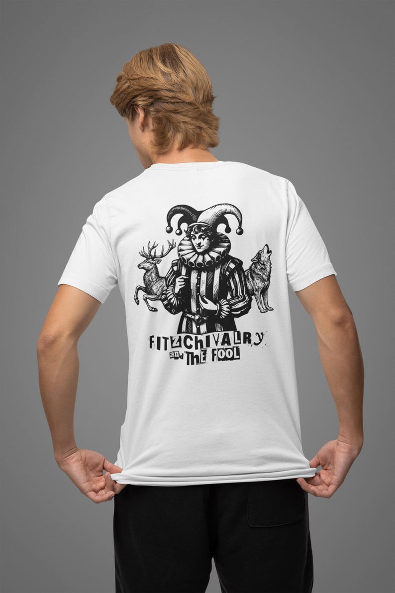 May include: White t-shirt with a black and white graphic of a jester figure, a deer, and a wolf. The text "FITUCHIVALRY and THE FOOL" is printed below the figures. The shirt is being worn by a person.