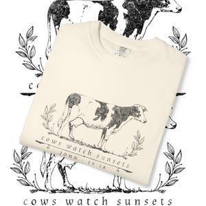 Pode incluir: Camiseta creme com uma ilustração de vaca em preto e branco e o texto "cows watch sunsets" e "JOHN 19:18". O design inclui detalhes florais e uma silhueta de vaca.