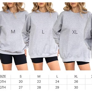 May include: Five women wearing gray sweatshirts in sizes S, M, L, XL, and 2XL. Each sweatshirt has a different size label. The women are wearing black shorts. A size chart below shows width and length measurements in inches.