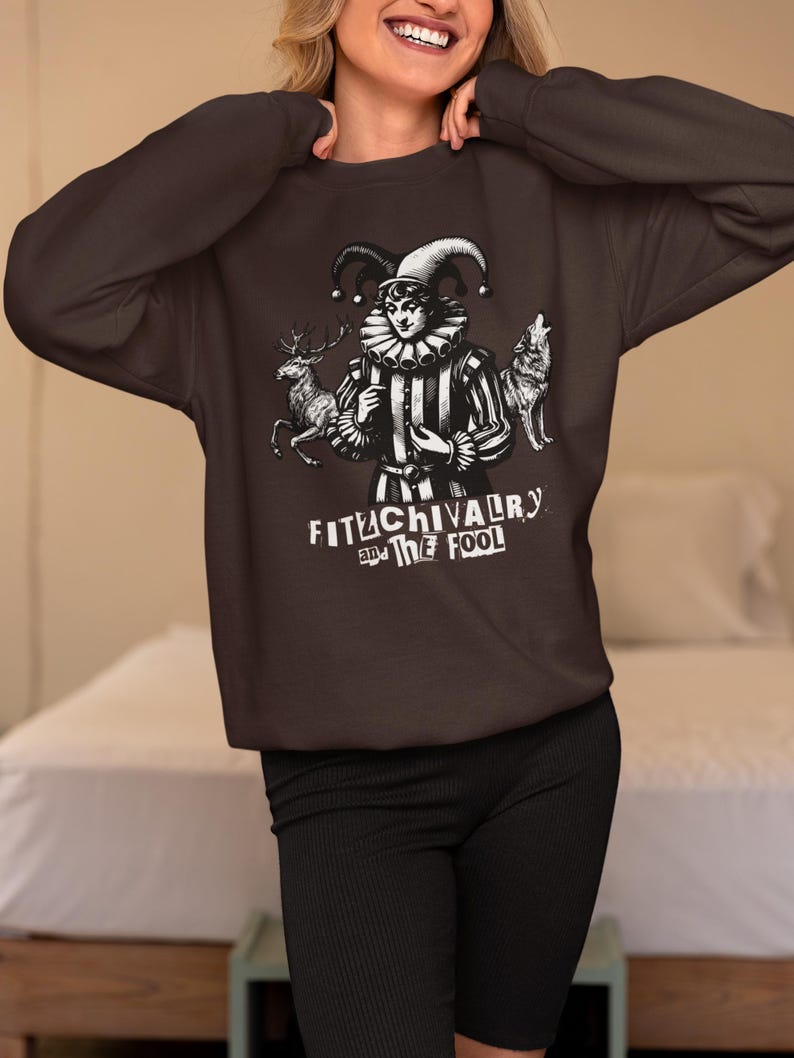 May include: A dark brown sweatshirt featuring a black and white graphic of a jester, deer, and wolf. The text "FITCHIVALRY and THE FOOL" is printed below the graphic. The sweatshirt is made of a soft material.