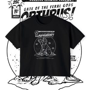 Könnte beinhalten: Ein schwarzes T-Shirt mit einem weißen Grafikdruck eines zweiköpfigen Hundes. Das Design enthält den Text "Dungeon Crawler Care #4", "Gate of the Feral Gods", "Orthrus!" und "The Universe's Goodest Boy" mit kleinen Sternen und Kreisdetails.
