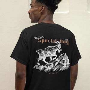 May include: Black t-shirt with a white graphic of a mountain goat leaping across a rocky landscape. Text above the graphic reads "Mommy's Special Boy" in a stylized font. The shirt is worn by a person.