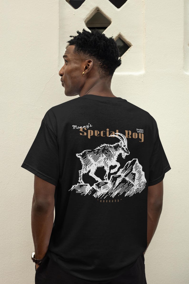 May include: Black t-shirt with a white graphic of a mountain goat leaping across a rocky landscape. Text above the graphic reads "Mommy's Special Boy" in a stylized font. The shirt is worn by a person.