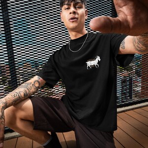 May include: Black t-shirt with a white goat graphic on the chest. The person is wearing black shorts and black boots. The background includes a metal fence and a blue sky.