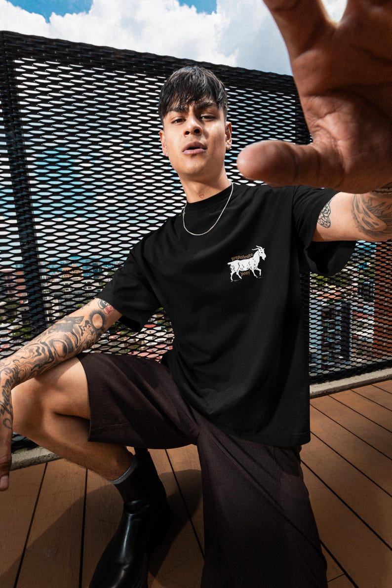 May include: Black t-shirt with a white goat graphic on the chest. The person is wearing black shorts and black boots. The background includes a metal fence and a blue sky.