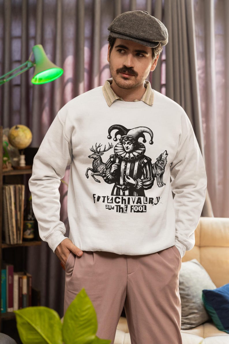 May include: White sweatshirt with a black and white graphic of a jester, deer, and wolf. The text "FITCHIVALRY and The Fool" is printed below the graphic. The person is wearing a gray cap and pink pants.
